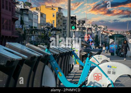 Fahrradverleih Fahrräder, die an der Fahrradstation geparkt sind. Ein Banner mit Anweisungen zum Ausleihen im Hintergrund und Menschen, die auf dem Gehweg laufen. Stockfoto