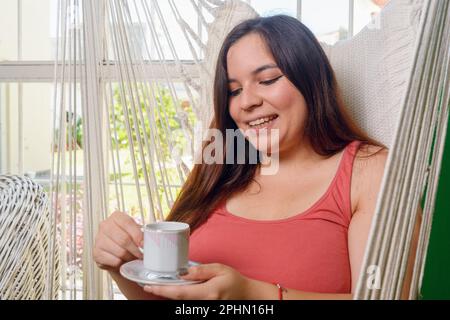 Porträt einer jungen latina, die zu Hause auf einem Stoffstuhl sitzt und Kaffee trinkt Stockfoto