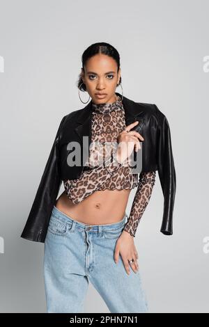 Stilvolles afroamerikanisches Modell in einem bauchfreien Oberteil mit Tiermuster und schwarzer Lederjacke isoliert auf Grau, Stockbild Stockfoto