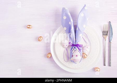Ostertisch. Weißer Teller mit einer Serviette, gefaltet in Form eines Kaninchens, Ostern und Schokoladeneiern auf pinkfarbenem Hintergrund. Frohe Osterferien Stockfoto