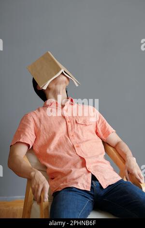 Ein junger Mann schlief auf einem Stuhl ein, mit einem geöffneten Buch im Gesicht Stockfoto