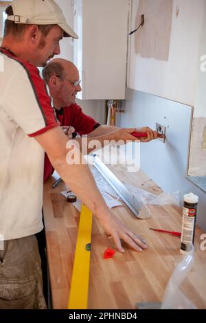 Kücheninstallateure: Elektrische Installation. Ein Elektriker zeigt seinem Auszubildenden, wie eine neue Steckdose (GB) eingebaut wird. Aus einer Reihe von zugehörigen Bildern. Stockfoto