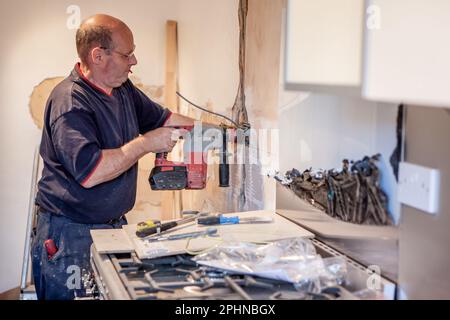 Kücheninstallateure: Elektrische Installation. Ein Elektriker, der Kabel in eine Wand bohrt, um eine neue Steckdose zu finden. Aus einer Reihe von zugehörigen Bildern. Stockfoto