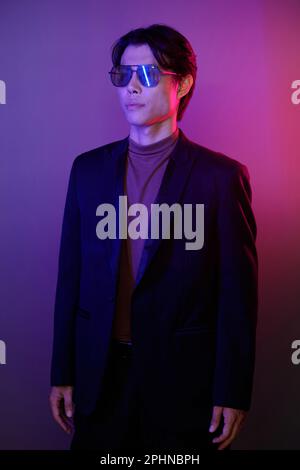 Porträt eines selbstbewussten jungen Mannes in Anzug und Sonnenbrille vor pinkfarbenem und lila Hintergrund Stockfoto