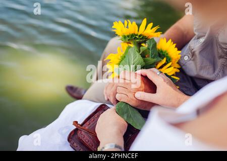 Älteres Familienpaar, das Händchen hält und sich am Sommerfluss entspannt. Eine Frau mit einem Strauß Sonnenblumen. Schließen. Romantisches Date Stockfoto
