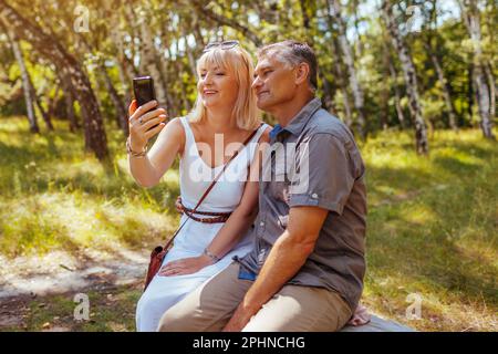 Porträt eines älteren Familienpaares, das Selfie mit dem Smartphone im Sommerwald macht. Ältere Menschen ruhen sich auf einer Bank aus und fotografieren. Mann und Frau Stockfoto