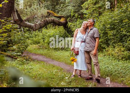 Stilvolles älteres Familienpaar, das im Sommerwald draußen spaziert. Ältere Menschen, die Händchen halten. Die Frau trägt einen Blumenstrauß. Mann und Frau genießen La Stockfoto
