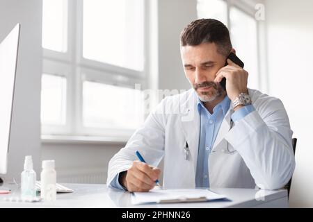Fokussierter männlicher Arzt, der mit dem Patienten telefoniert und in das Klemmbrett schreibt, Notizen macht, in der Klinikpraxis arbeitet Stockfoto