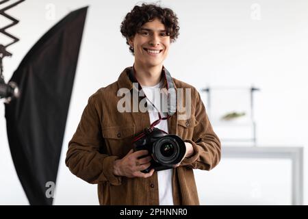 Glücklicher junger Fotograf mit professioneller Kamera, arbeitet im modernen Fotostudio, lächelt vor der Kamera Stockfoto
