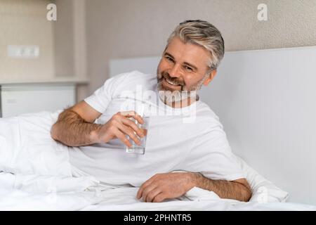 Glücklicher, reifer, weißer Mann, der ein Glas Wasser hält, Schlafzimmer-Inneneinrichtung Stockfoto