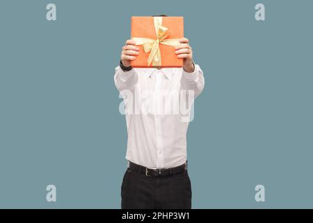 Porträt eines unbekannten anonymen Mannes, der sein Gesicht mit einer roten Geschenkbox überdeckt, ein Geschenk zum Jubiläum verschenkt, ein Indoor Studio mit isoliertem Bild auf hellblauem Hintergrund trägt. Stockfoto