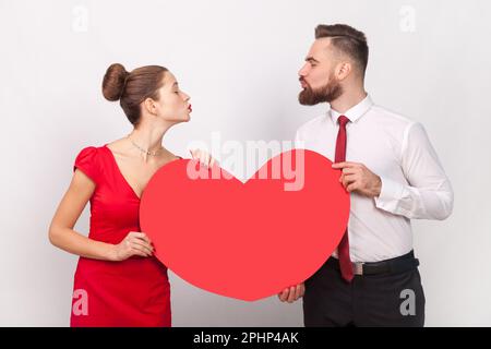 Portrait eines reizenden Mannes im Hemd und einer Frau im roten Kleid, die zusammenstehen, sich Luftküsse zuschicken, ein großes Herz in der Hand halten. Studioaufnahmen im Innenbereich isoliert auf grauem Hintergrund. Stockfoto