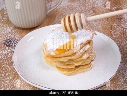 Hausgemachter Stapel Pfannkuchen auf einem weißen Teller mit einer Tasse Kaffee und Honig auf einem Holztisch, Bild-ID: 2PHP8HK Stockfoto