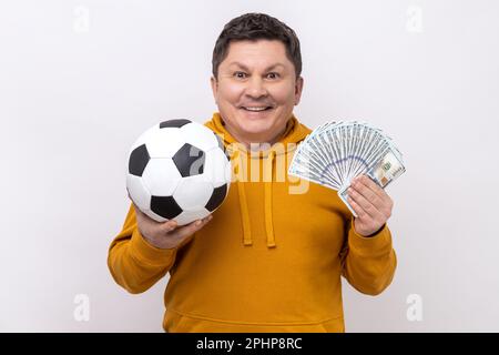 Ein lächelnder Mann mittleren Alters, der Fußball zeigt und Hundert-Dollar-Scheine spielt, viel Geld beim Sport gewinnt, einen urbanen Hoodie trägt. Studioaufnahmen im Innenbereich isoliert auf weißem Hintergrund. Stockfoto