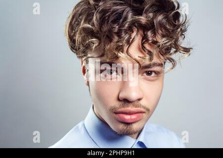 Porträt eines gutaussehenden Mannes mit Schnurrbart und Bart mit lockiger Frisur, mit blauem Hemd, der mit ernstem Gesichtsausdruck in die Kamera schaut. Studioaufnahmen im Innenbereich isoliert auf grauem Hintergrund. Stockfoto