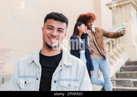Vorderansicht eines jungen, gut aussehenden arabischen Mannes, der im Freien lächelt und steht, im Hintergrund, während seine beiden multiethnischen Freunde ein Selfie-Porträt zusammen machen. Ein glücklicher Teenager, der in die Kamera schaut. Hochwertiges Foto Stockfoto