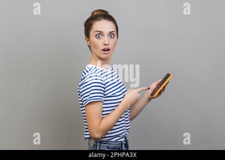 Porträt einer erstaunten Bloggerin, die ein gestreiftes T-Shirt am Smartphone trägt, mit offenem Mund auf die Kamera schaut und schockierende Inhalte sieht. Studioaufnahmen im Innenbereich isoliert auf grauem Hintergrund. Stockfoto