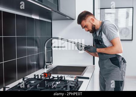 Junger professioneller Klempner in grauer Uniform, der den Wasserhahn in der Küche fixiert Stockfoto