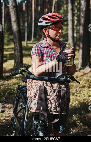 Aktive Frau verbringt kostenlose Sommerferien auf einer Fahrradtour in einem Wald. Frau mit Fahrradhelm und Handschuhen, die das Fahrrad hält, mit Korb und dr. Stockfoto