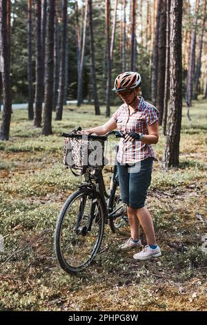Aktive Frau, die freie Sommerferienzeit auf einer Fahrradtour in einem Wald verbringt. Frau trägt Fahrradhelm und Handschuhe halten Fahrrad mit Korb Stockfoto