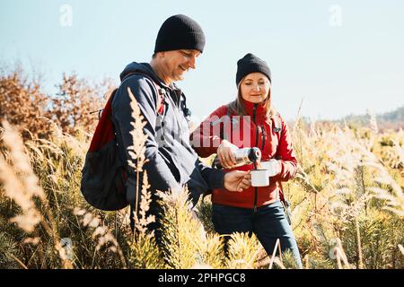 Paare entspannen und genießen den Kaffee während des Urlaubs. Leute stehen auf dem Weg und gießen Kaffee aus Thermoskanne. Paar mit Rucksäcken wandern Stockfoto