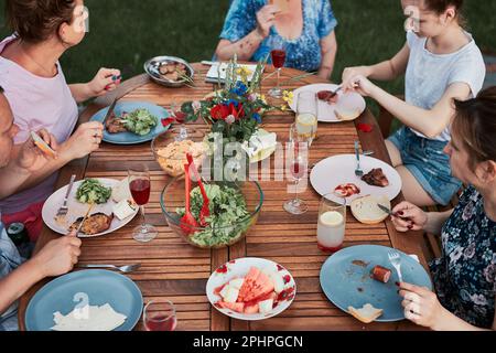 Familie mit einer Mahlzeit vom Grill während des Sommer Picknick im Freien Abendessen in einem Hausgarten. Nahaufnahme von Leuten, die an einem Tisch mit Essen und Geschirr sitzen Stockfoto