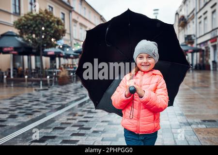 Wenig lächelnd glücklich Mädchen hält großen Regenschirm zu Fuß in einem Innenstadt an regnerisch düsteren Herbsttag Stockfoto