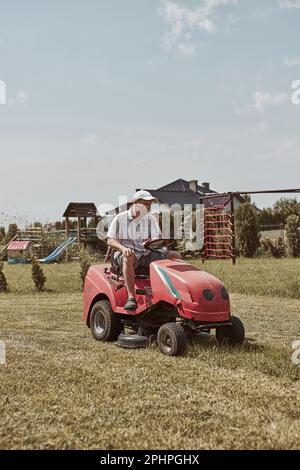 Mann mäht seinen Rasen mit Rasenmäher fahren. Mann, der Gras in seinem Garten neben dem Haus beibehält Stockfoto