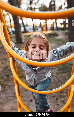 Verspielte, glückliche Vorschulkinder, die auf einem Spielplatz spielt, auf Affenbars klettert und mit geschlossenen Augen lächelt. Kind hat Spaß beim Spielen im Freien in einem Park o Stockfoto