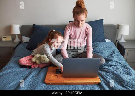 Teenager Mädchen, die an der Schule arbeiten, testet Hausaufgaben von zu Hause aus auf ihrem Laptop während der COVID-19-Quarantäne in Begleitung ihrer kleinen Schwester. Mädchen Sitti Stockfoto