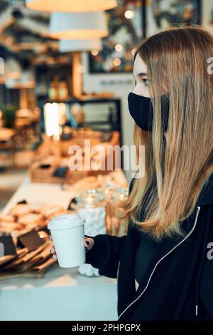 Eine Frau, die eine Kaffeetasse in der Hand hält, in einem Café in der Schlange steht und auf die Bestellung wartet. Person, die die Gesichtsmaske trägt, um Virusinfektion zu vermeiden und p Stockfoto