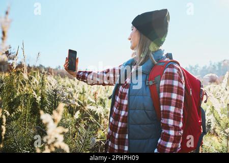 Junge Frau macht Selfie mit Smartphone während einer Bergreise. Frau mit Rucksack wandert durch hohes Gras auf der Wiese. Ausgaben Stockfoto