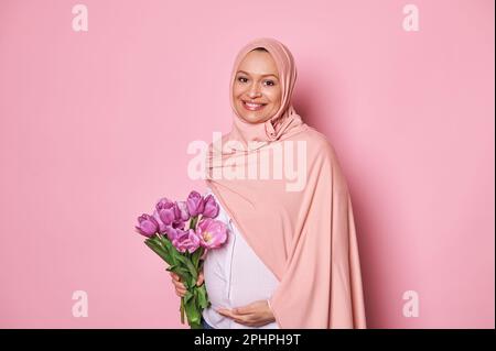 Muslimische Frau in pinkem Hijab, hält einen Strauß Tulpen in der Hand, berührt ihren schwangeren Bauch und ist glücklich, wenn das Baby zuerst tritt Stockfoto
