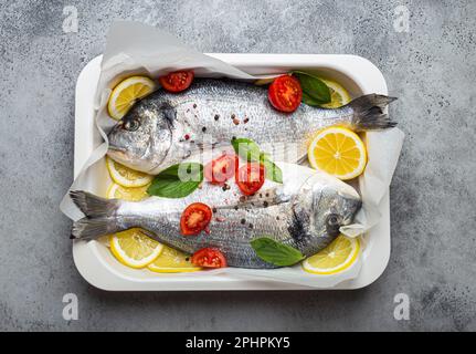 Zwei rohe Fische dorado in einem Auflauf mit Zutaten wie Zitrone, frischem Basilikum und Kirschtomaten aus nächster Nähe auf rustikalem steingrauem Hintergrund Stockfoto