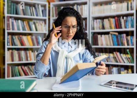 Müde, überarbeitete Studentin, die allein in der Universitätsbibliothek studiert, Frau, die kein komplexes Material versteht, hart lernt und sich auf die Prüfung vorbereitet, hispanische Frau mit lockigem Haar und Brille. Stockfoto