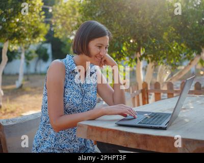 Glückliche junge asiatische Frau mit Laptop, die sich draußen in einem winzigen Haus im Sommergarten ausruht, Wochenendausflug und eine Idee für ein abgelegenes Büro, am frühen Morgen. Winzige Häuser A Stockfoto