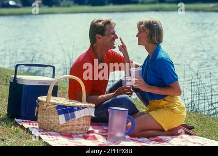 1990 historische junge Paar PICKNICK AUF DECKE DURCH WASSER Stockfoto