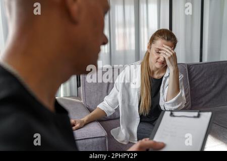 Ein männlicher Psychologe konsultiert eine Patientin, macht Notizen. Eine Frau spricht emotional über sich selbst, bedeckt ihr Gesicht mit ihren Händen. Stockfoto