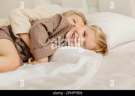 Die Brüder liegen in Morgenmänteln in einem weißen Bett, umarmen, lächeln. Familienkonzept. Stockfoto