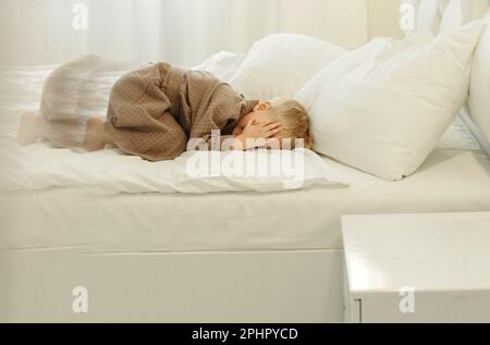 Ein Junge im braunen Bademantel, liegt auf einem weißen Bett, bedeckt sein Gesicht mit den Händen. Hinter der Reflexion. Stockfoto