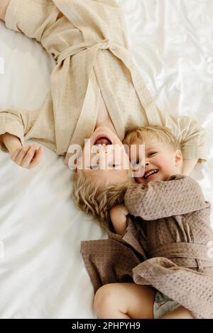 Die Brüder liegen in Morgenmänteln in einem weißen Bett, spielen, umarmen, lächeln. Familienkonzept. Blick von oben. Stockfoto