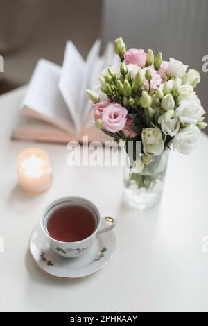 Ein Stillleben im Frühling, ein Strauß Rosen in einer Vase, eine Tasse Tee und ein Buch auf einem weißen Tisch. Frühlingsgrußkarte mit Frühlingsblumen. Romantisch Stockfoto
