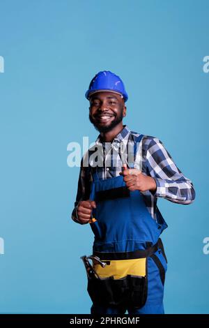 Aufgeregter afroamerikanischer Mann mit Schutzhelm zeigt Daumen hoch als Zeichen der Anerkennung. Optimistischer Baumeister, der mit Werkzeugen und Uniform, Bauarbeiten und Renovierungen einen Gürtel trägt. Stockfoto