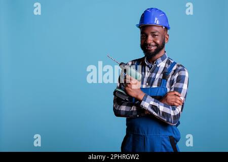 Ein afroamerikanischer Baumeister, der einen elektrischen Akku-Bohrer hält, der glücklich im Studio sitzt. Konstrukteur trägt Schutzhelm und Overall, Sicherheitsuniform im Baugewerbe. Stockfoto