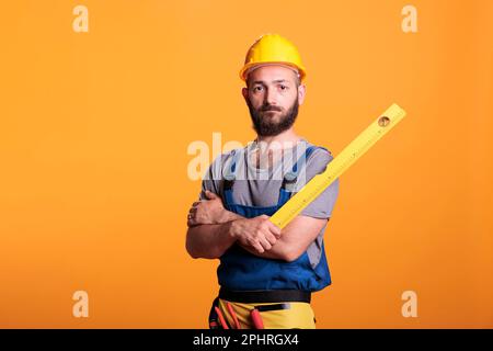 Bauarbeiter, der das Wasserstandlineal im Studio hält, posiert mit gekreuzten Armen. Junger Mechaniker, der sich auf Gebäudemessungen vorbereitet, mit einem professionellen Nivellierwerkzeug, einem selbstbewussten Zimmermann. Stockfoto