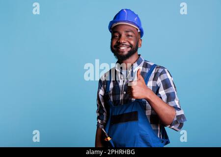 Stolzer afroamerikanischer professioneller Elektriker mit Overalls und Schutzhelm. Zeigt Daumen nach oben und freut sich über seine Arbeit, während er vor blauem Hintergrund lächelt. Hintergrund. Stockfoto