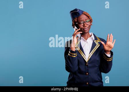 Wütende weibliche Flugbegleiterin, die telefoniert, in Uniform gekleidet, mit Händen gestrickt. Stewardess wirkt verärgert, wenn sie im Studio ein Mobilgerät verwendet, das vor blauem Hintergrund aufgenommen wurde. Stockfoto