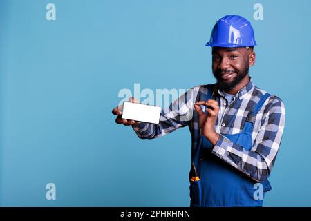 Mann in Arbeitsuniform hält Smartphone mit leerem Werbeleitschirm in Studioaufnahme. Ein afroamerikanischer Bauarbeiter zeigt auf einen leeren Handybildschirm auf blauem Hintergrund. Stockfoto