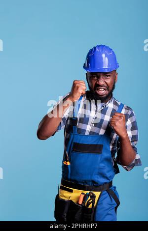 afroamerikanische männliche Fäuste, um vor der Kamera zu schlagen, drücken wütende Reaktionen aus, die sich auf einen Kampf vorbereiten. Verärgerter Bauarbeiter, der sich in den Kampf stürzte, Studioaufnahme vor blauem Hintergrund. Stockfoto