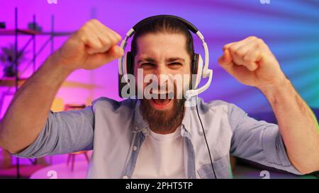 Ein bärtiger Gamer in einem Gaming-Headset gewinnt ein Videospiel. Cybersportler in einem Neonraum. Lifestyle-Gamer, Online-Spiele. Sieg im E-Sport Stockfoto